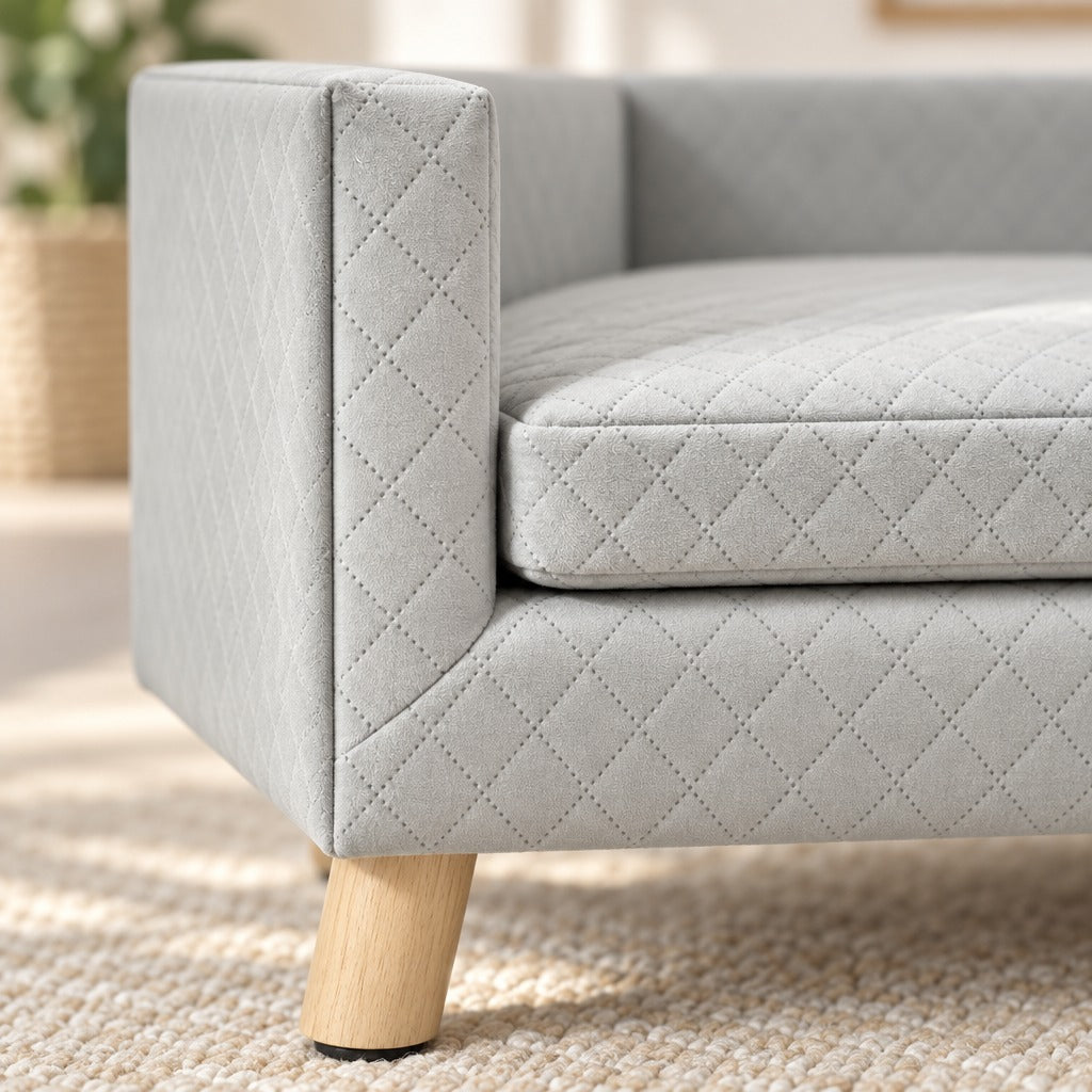 Gros plan sur l'angle avant de la banquette gris clair : velours matelassé de losanges, accoudoir et pied en bois clair à patin antidérapant.