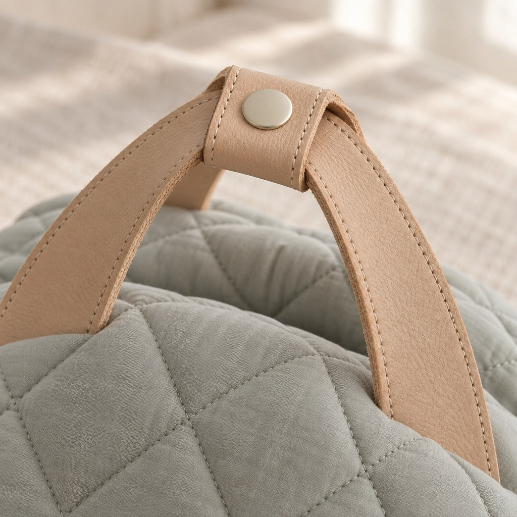 Gros plan sur le passant de cuir à bouton pression qui réunit les deux anses cousues au sommet de la coque matelassée gris sauge pâle.