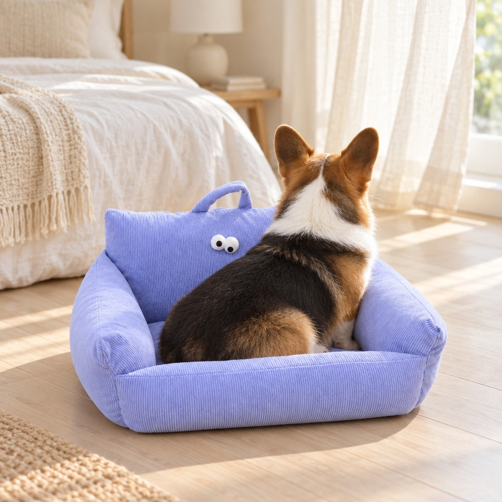 Corgi tricolore assis de dos dans le couchage canape en velours cotele lavande, au pied d'un lit clair.