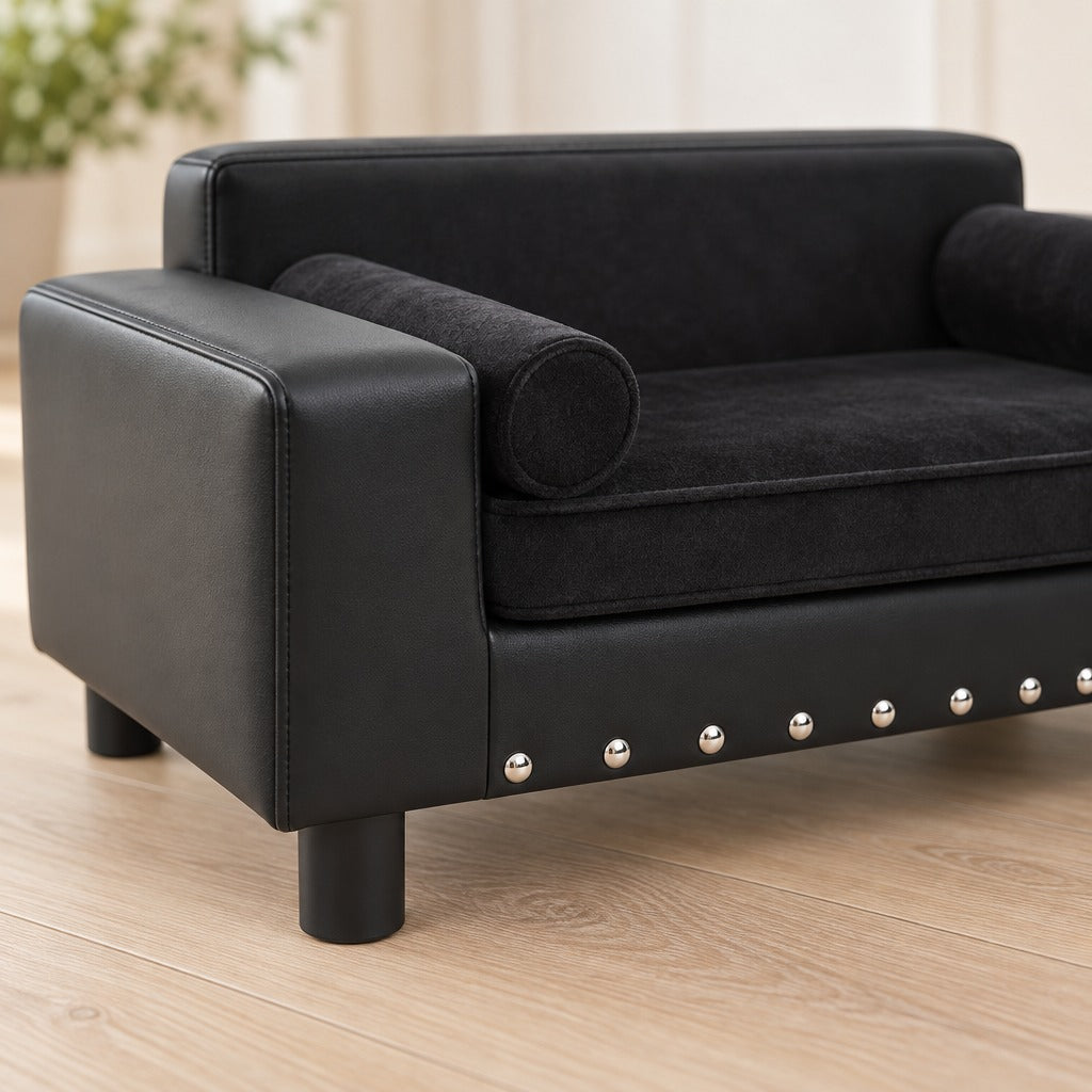 Gros plan sur l'angle avant du canapé noir en simili, son bandeau clouté, un pied cylindrique et la peluche mate du coussin.