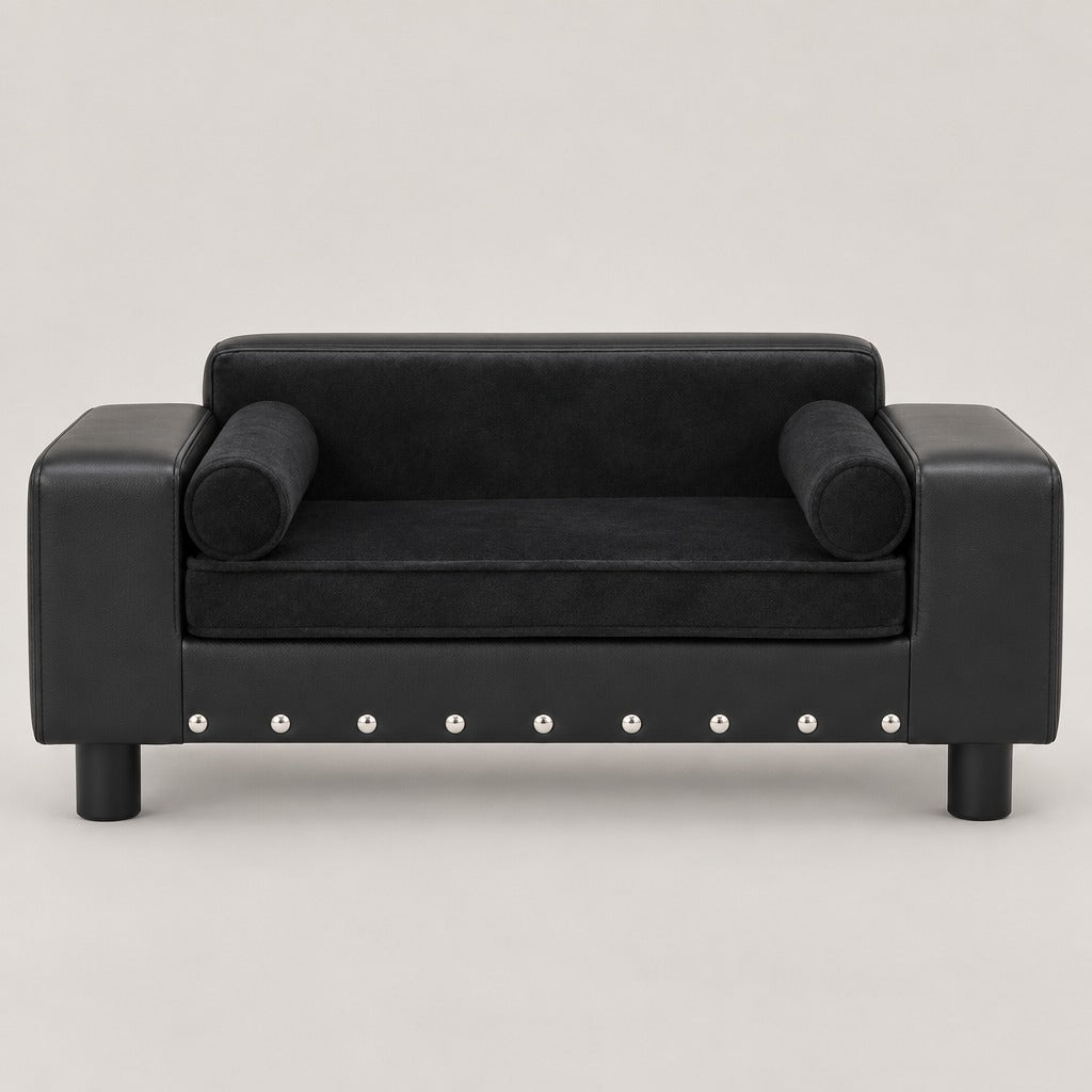 Canapé noir en simili vu de face, ses deux traversins en peluche posés contre les accoudoirs et son bandeau avant clouté.