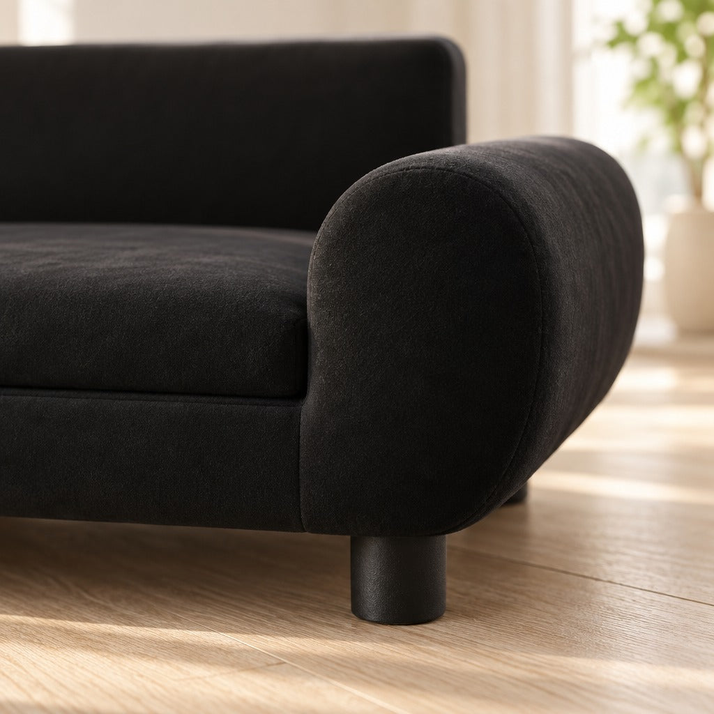 Gros plan sur l'accoudoir roulé en velours noir, le grain du tissu bien lisible et un pied rond noir posé sur le parquet clair.