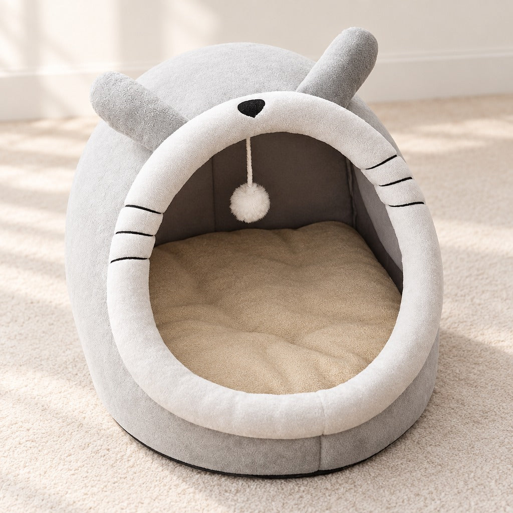 Cocon pour chat gris perle vu de trois quarts, ouverture frontale, coussin sable et pompon suspendu.