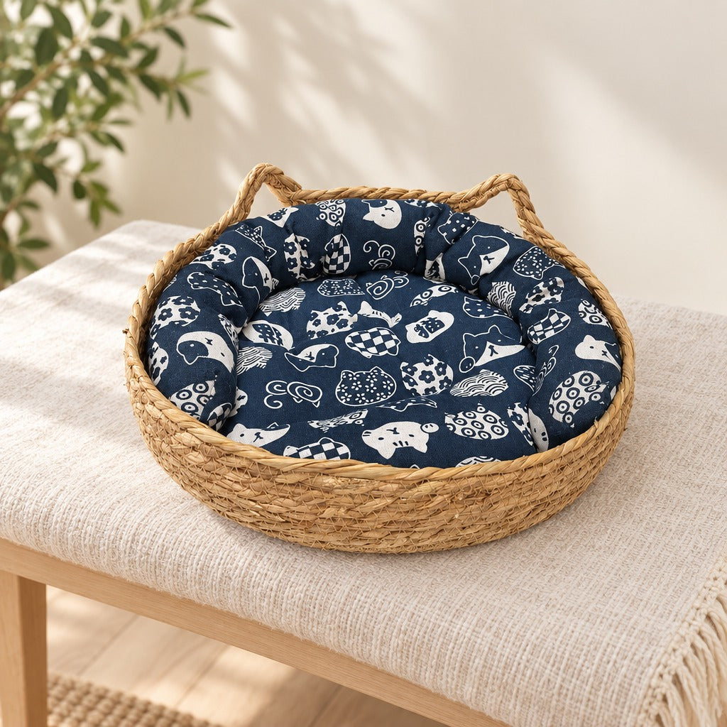 Corbeille pour chat en fibre végétale tressée posée seule sur un banc clair, avec son coussin bleu marine imprimé chats et poissons.