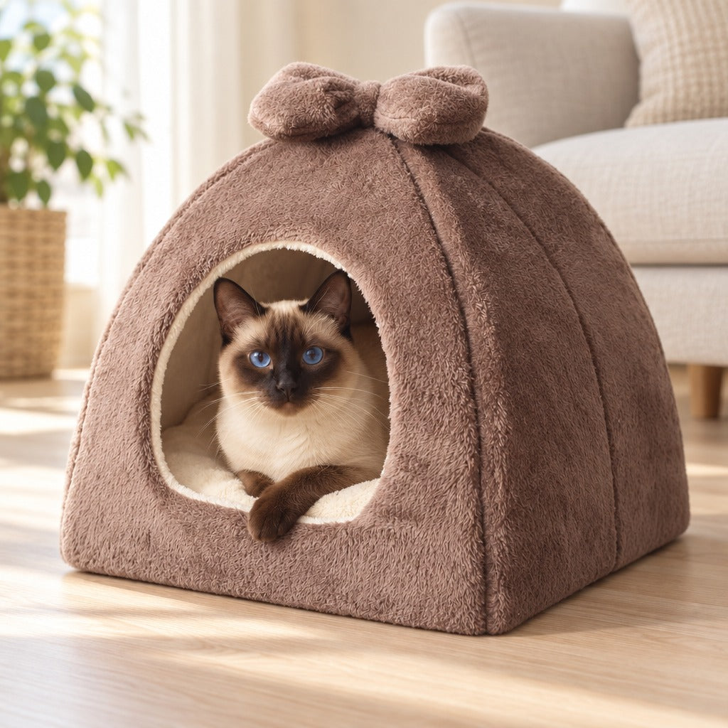 Un chat siamois seal point installé dans l'ouverture du dôme en peluche brun cacao, une patte avant posée sur le bord, dans un salon clair.