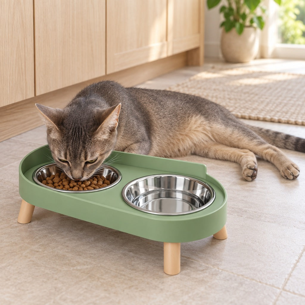 Chat tigré gris allongé qui mange dans la gamelle double surélevée vert sauge posée dans une cuisine claire.
