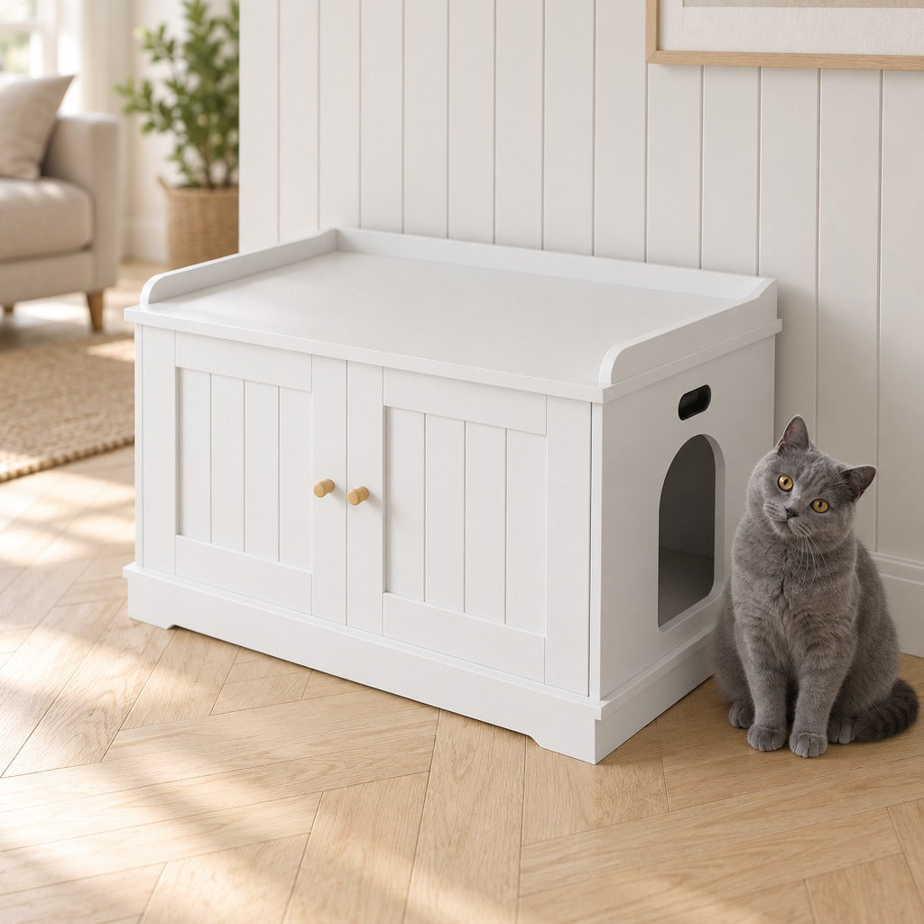 Chat gris bleu assis à côté du meuble à litière coloris Blanc, tout près de l'entrée arrondie de son flanc.