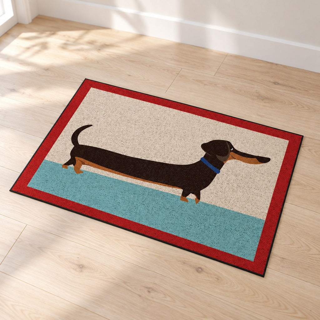 Le paillasson crème, turquoise et rouge seul sur un parquet clair, vu de trois quarts, son teckel imprimé lisible d'un bout à l'autre.