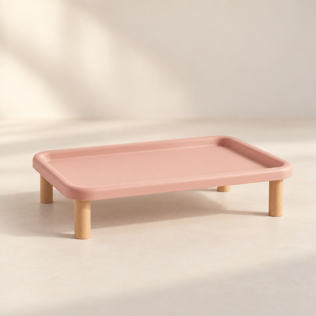 Le plateau repas surélevé rose poudré seul et vide sur fond clair, vu de trois quarts, bord relevé continu et quatre pieds en bois.