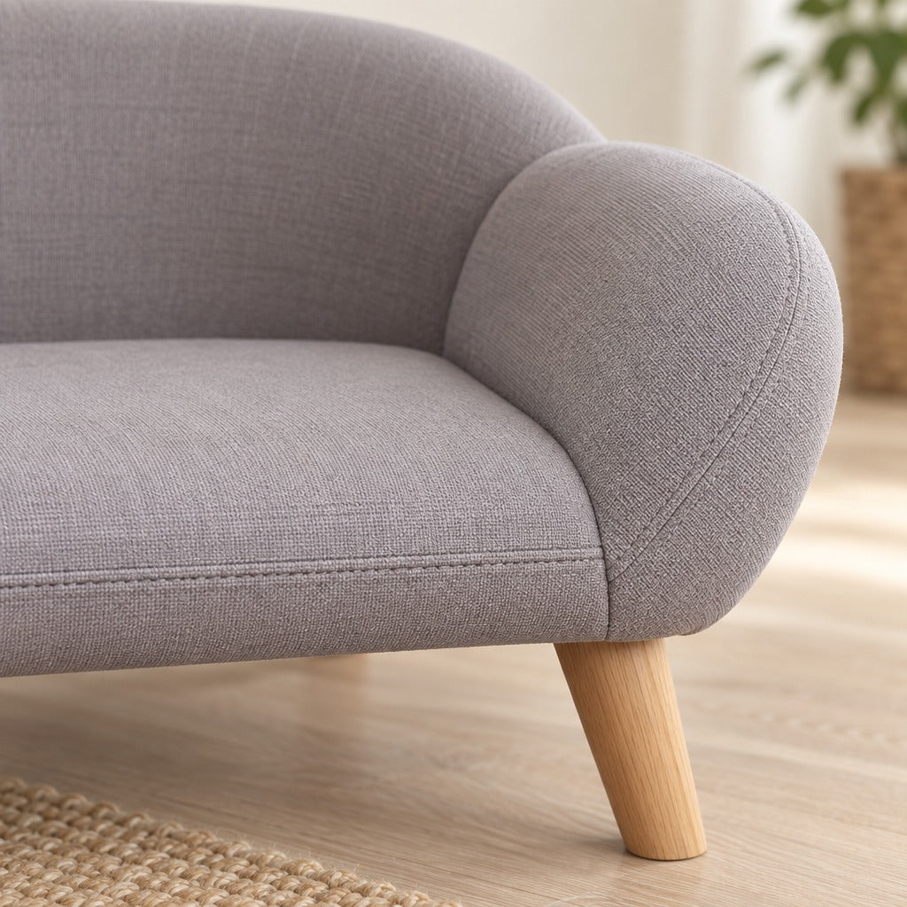Gros plan sur le tissu tisse gris clair de l'accoudoir arrondi et sur la surpiqure du bord d'assise, avec le haut d'un pied en bois clair.