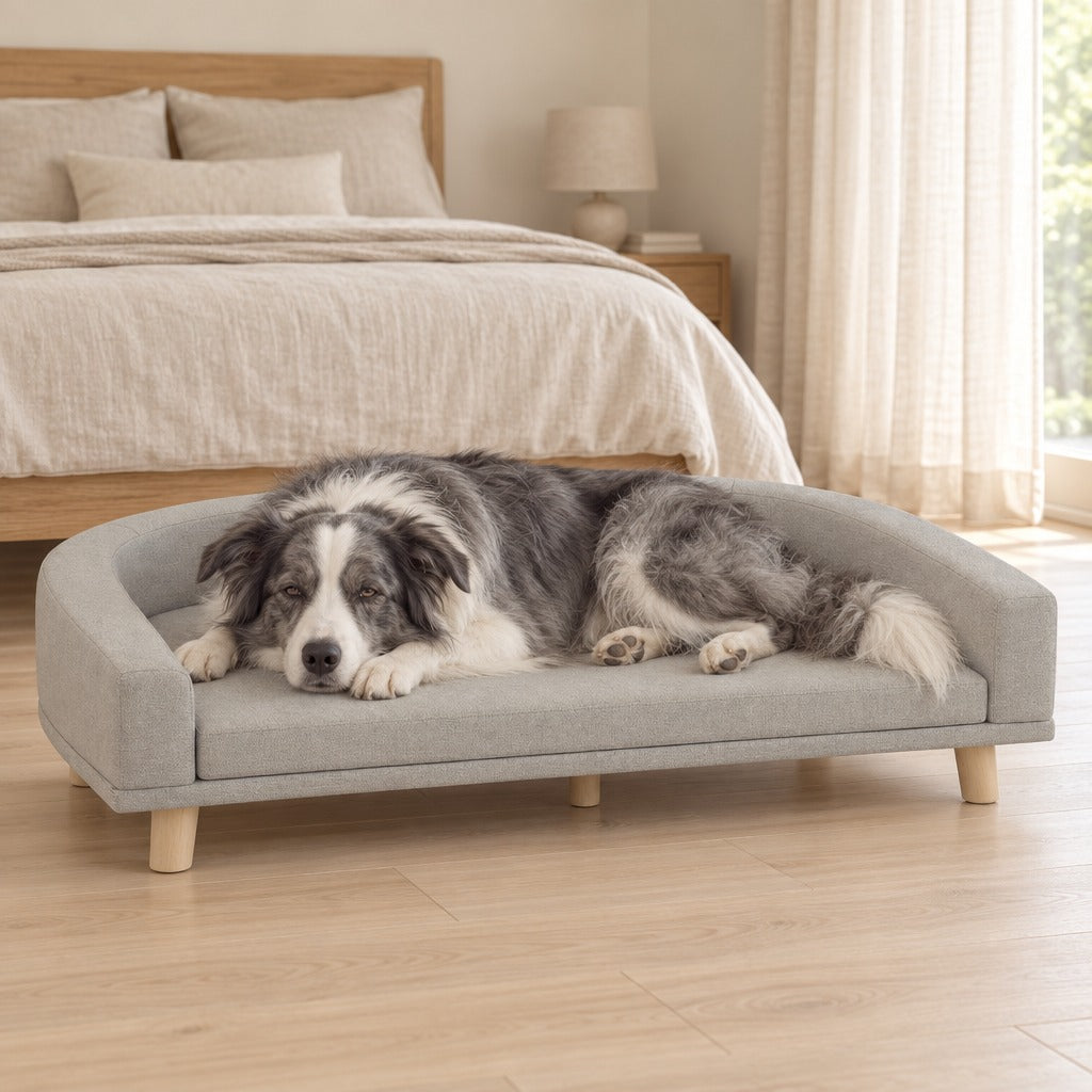 Un border collie gris et blanc assoupi sur le canapé pour chien gris clair installé au pied d'un lit habillé de lin.