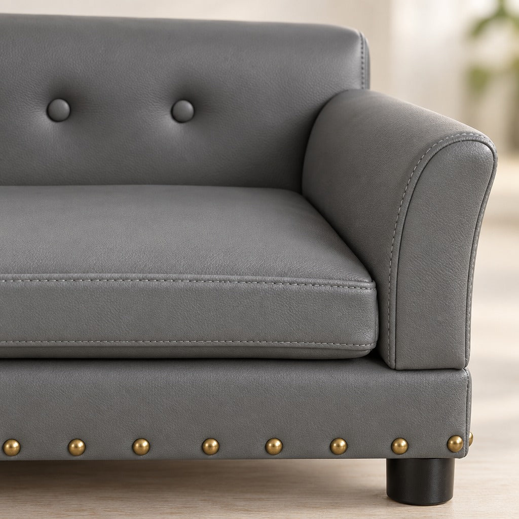 Gros plan sur le similicuir gris du sofa rectangulaire, un bouton de capitonnage et la rangée de clous dorés du socle.