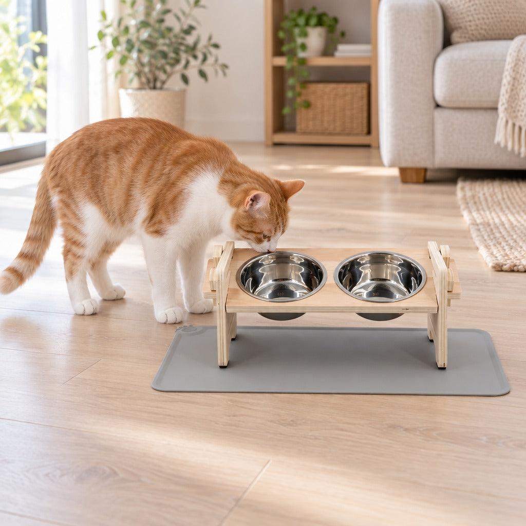 Chat blanc et roux qui renifle le bord du support de gamelles en bois clair, coloris bois clair et gris, posé sur son tapis gris dans un salon lumineux.
