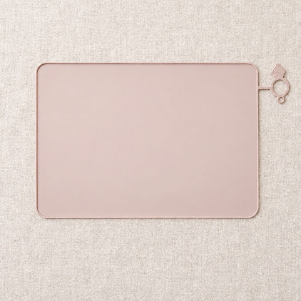 Tapis de gamelle pour chat rose poudré vu de dessus, seul sur une toile de lin claire, avec sa patte d'accroche à anneau.