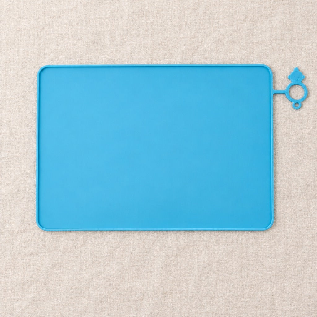 Tapis de gamelle pour chat bleu ciel vu de dessus, seul sur une toile de lin claire, avec sa patte d'accroche à anneau.